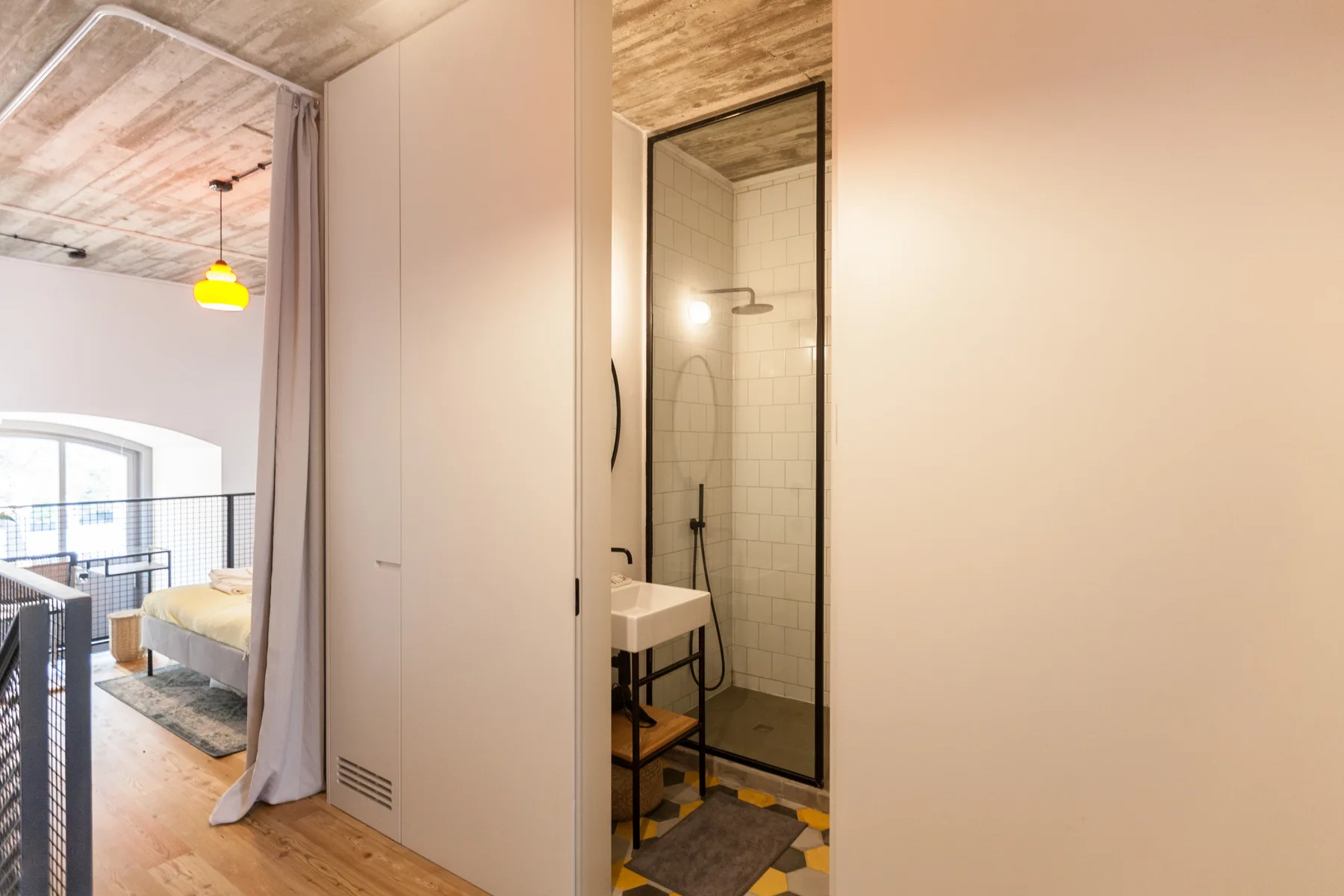 Un interior compacto muestra una ducha alicatada junto a un dormitorio con cama y asientos.