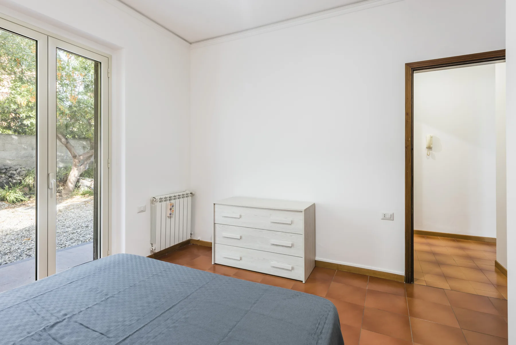 Un dormitorio con una cama, una cómoda pequeña y una puerta de vidrio que da a un jardín.