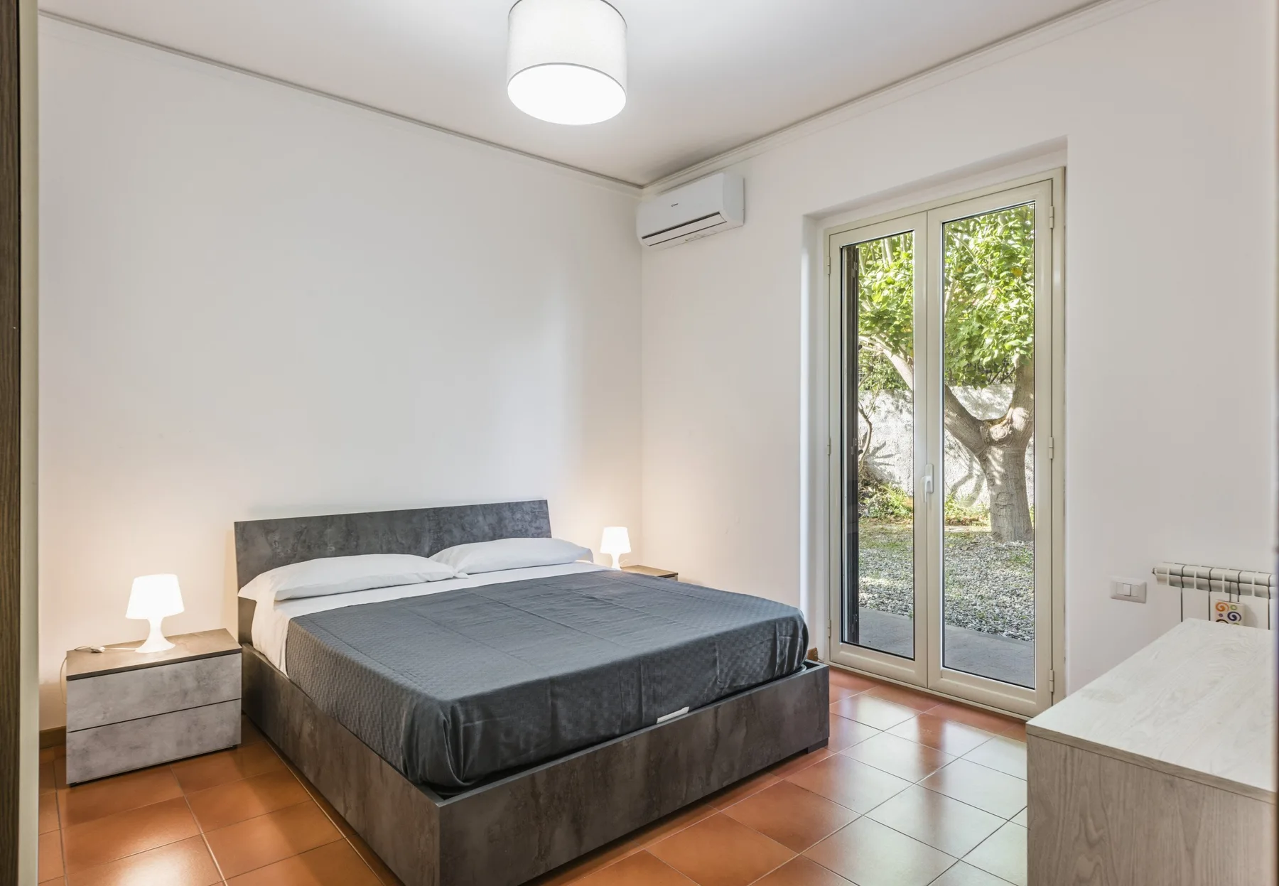 Dormitorio con una cama, mesitas de noche y una puerta acristalada que da a la vegetación.