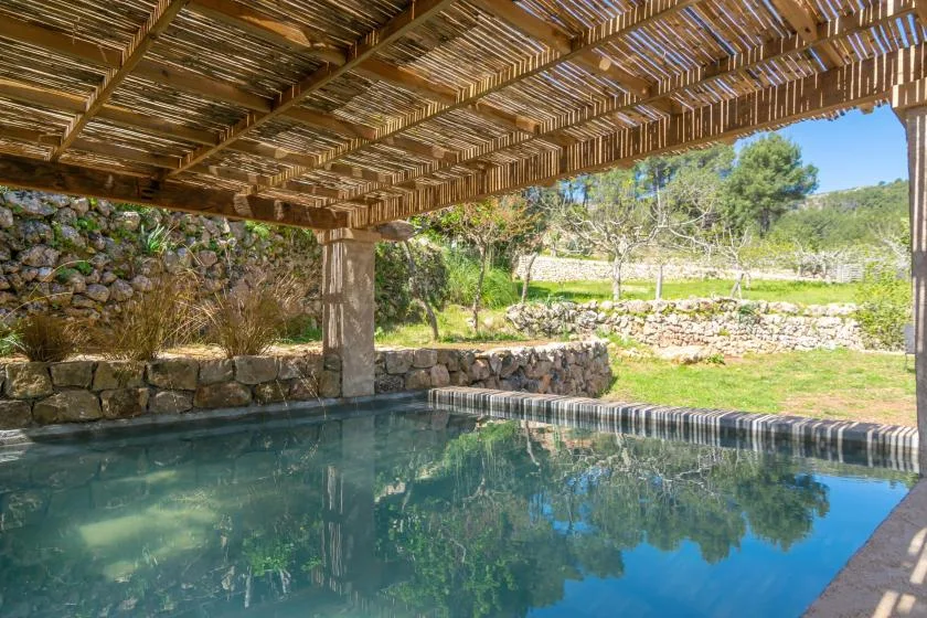 Una piscina está bajo una pérgola rústica, con muros de piedra y vegetación al fondo.