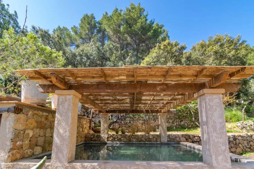 Una piscina exterior está bajo una pérgola de madera, rodeada de árboles y muros de piedra.