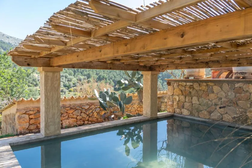 Una piscina al aire libre está bajo una pérgola de madera, junto a muros de piedra y una ladera.