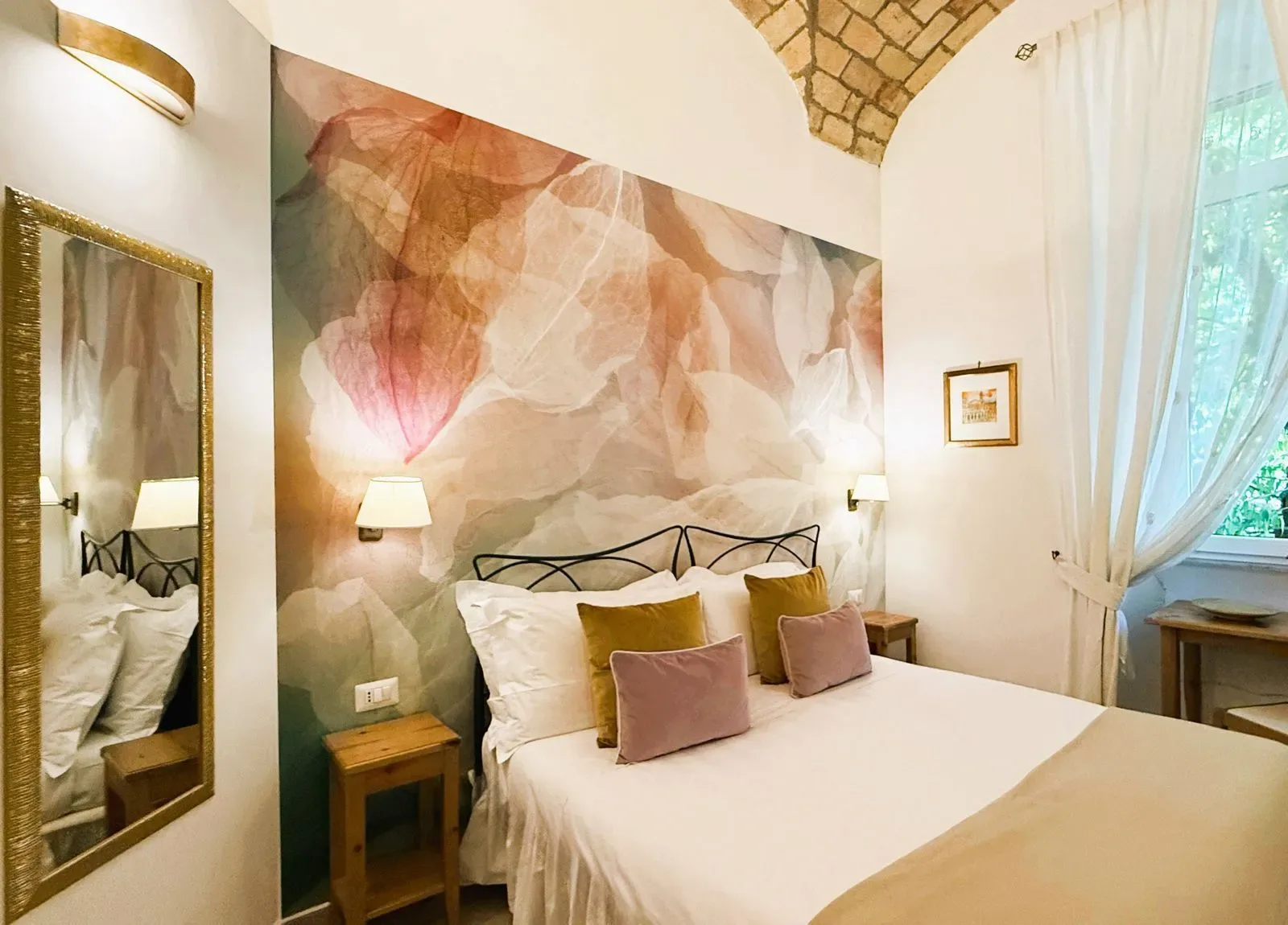 Dormitorio con mural floral, cama, techo abovedado, espejo y ventana con cortinas.