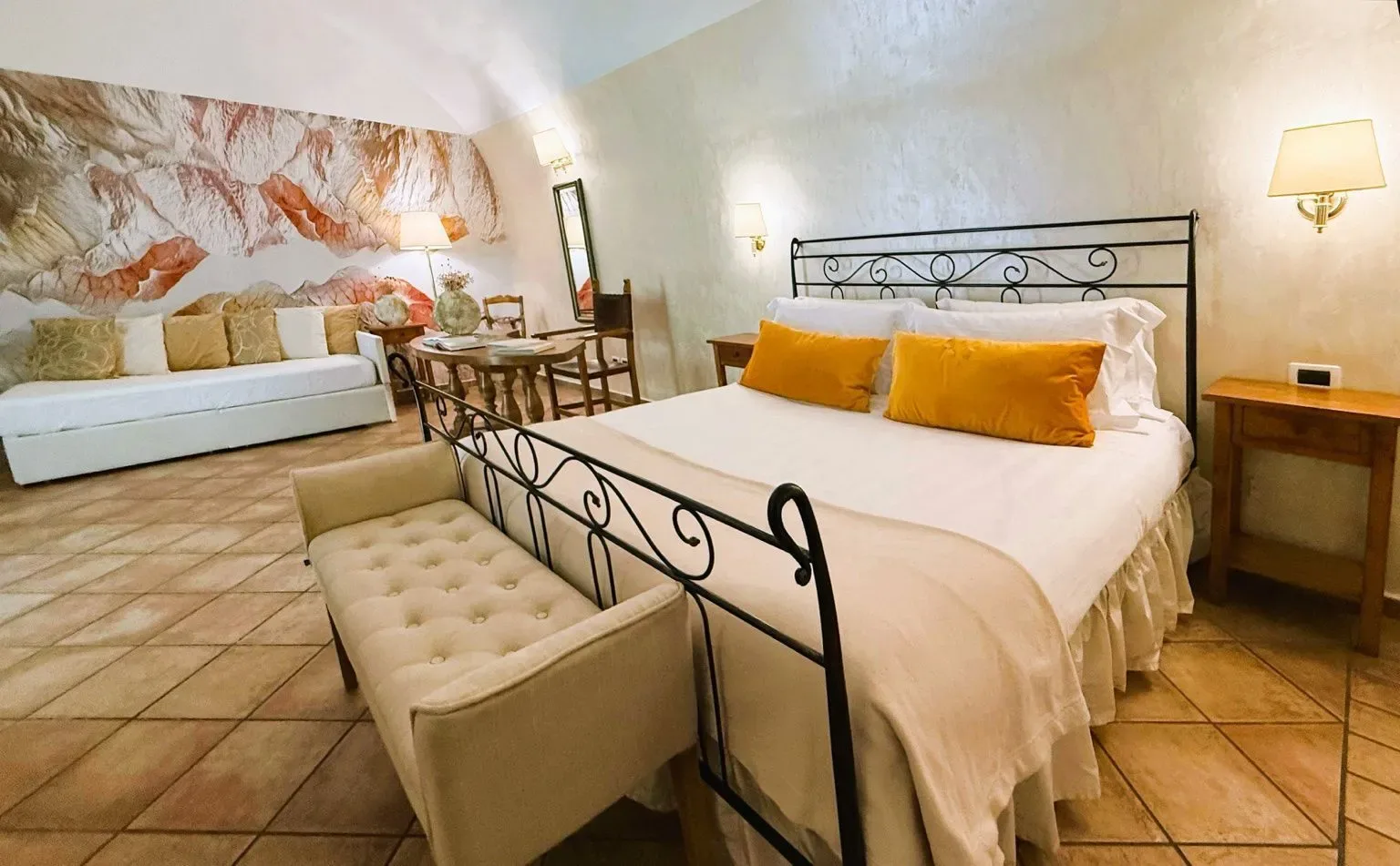 Dormitorio con cama de hierro forjado, diván, mesa y suelo de madera, y mural floral.