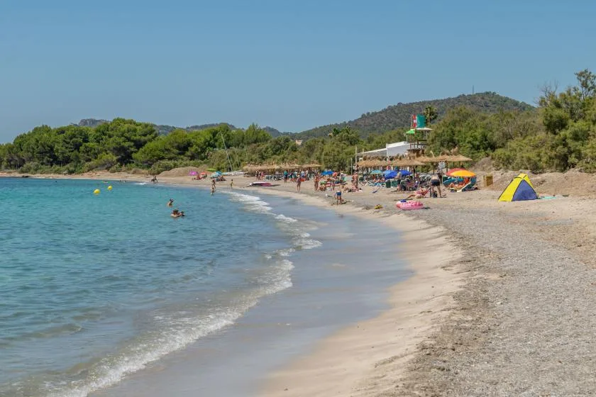 A beach curves along clear water, with beachgoers and colorful umbrellas on the shore.