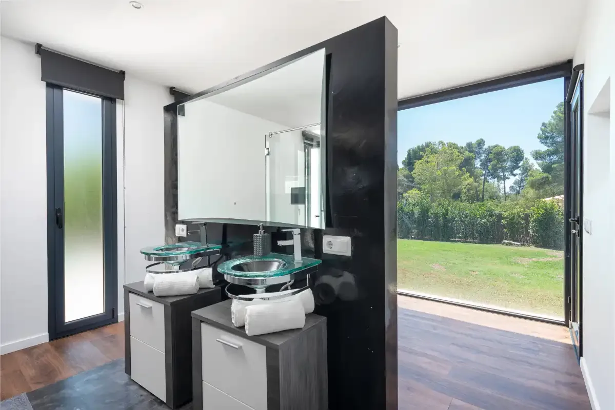 A modern bathroom with glass sinks beside a large window overlooking a green yard.