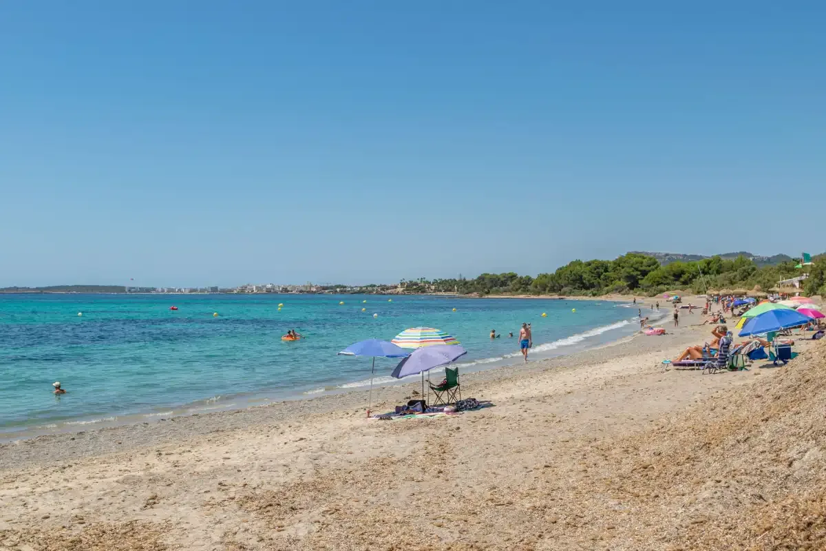 A sandy beach borders clear blue water, with umbrellas and people along the shore.