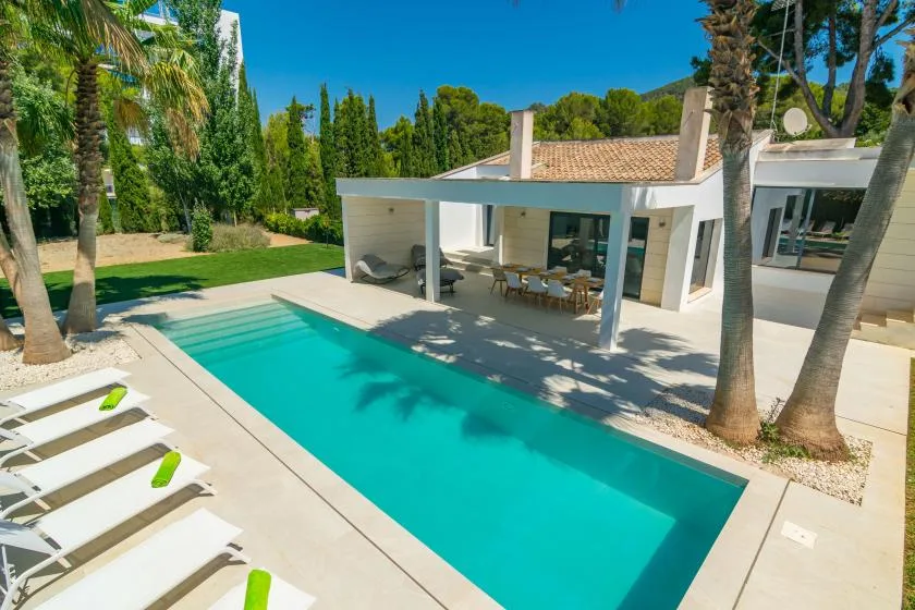 An outdoor swimming pool beside a modern house, with lounge seating on the patio.