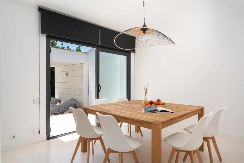 A bright dining area with a wooden table, white seating, and a large glass door.