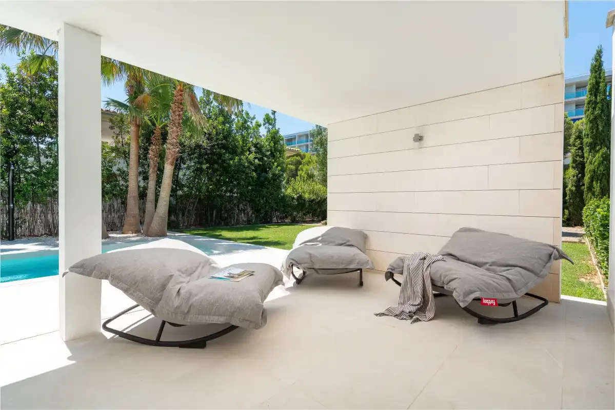 A covered outdoor patio with cushioned lounge seating overlooks a garden and pool.