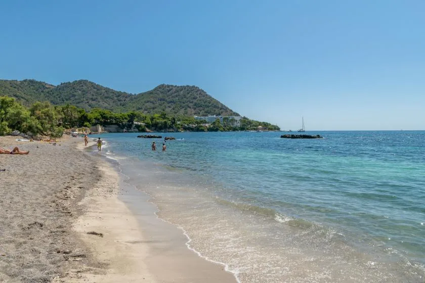 A sandy beach curves along clear blue water beneath green hills.