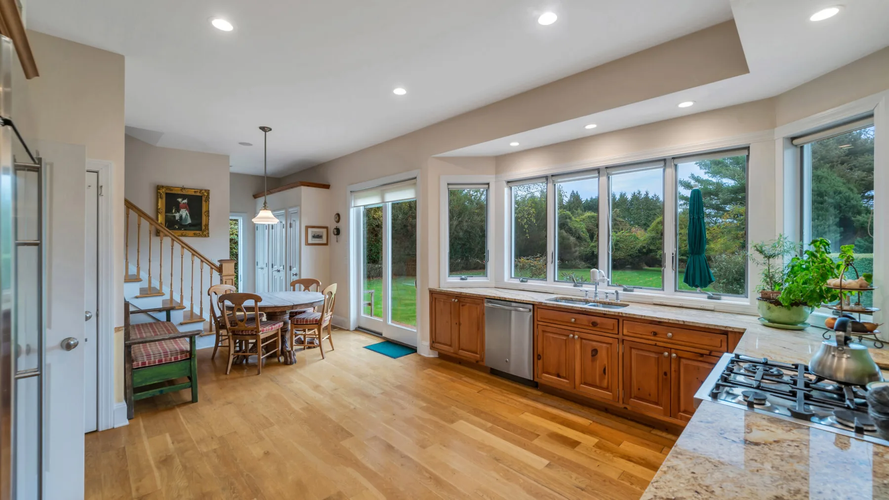 Una cocina y comedor luminosos tienen gabinetes de madera, ventanas grandes y una mesa de comedor.