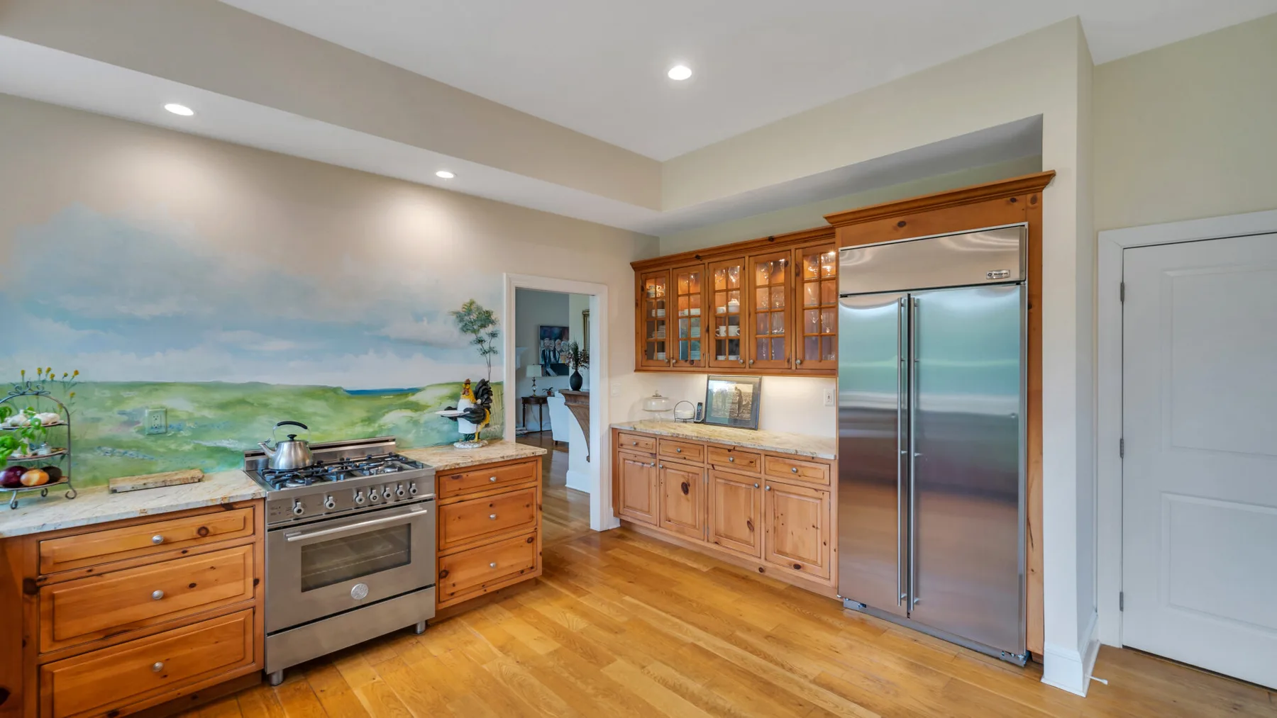 Una cocina amplia con gabinetes de madera, un refrigerador de acero inoxidable y un mural de paisaje.