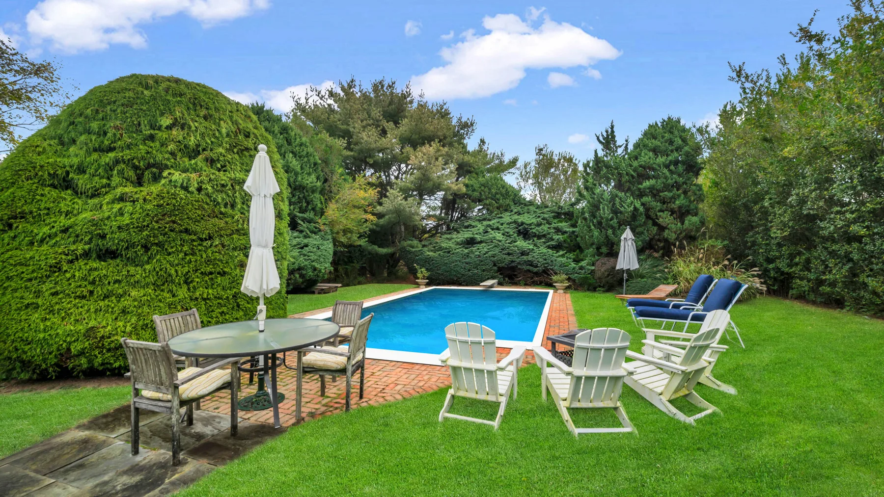 An outdoor swimming pool sits beside a lawn with patio seating and umbrellas.