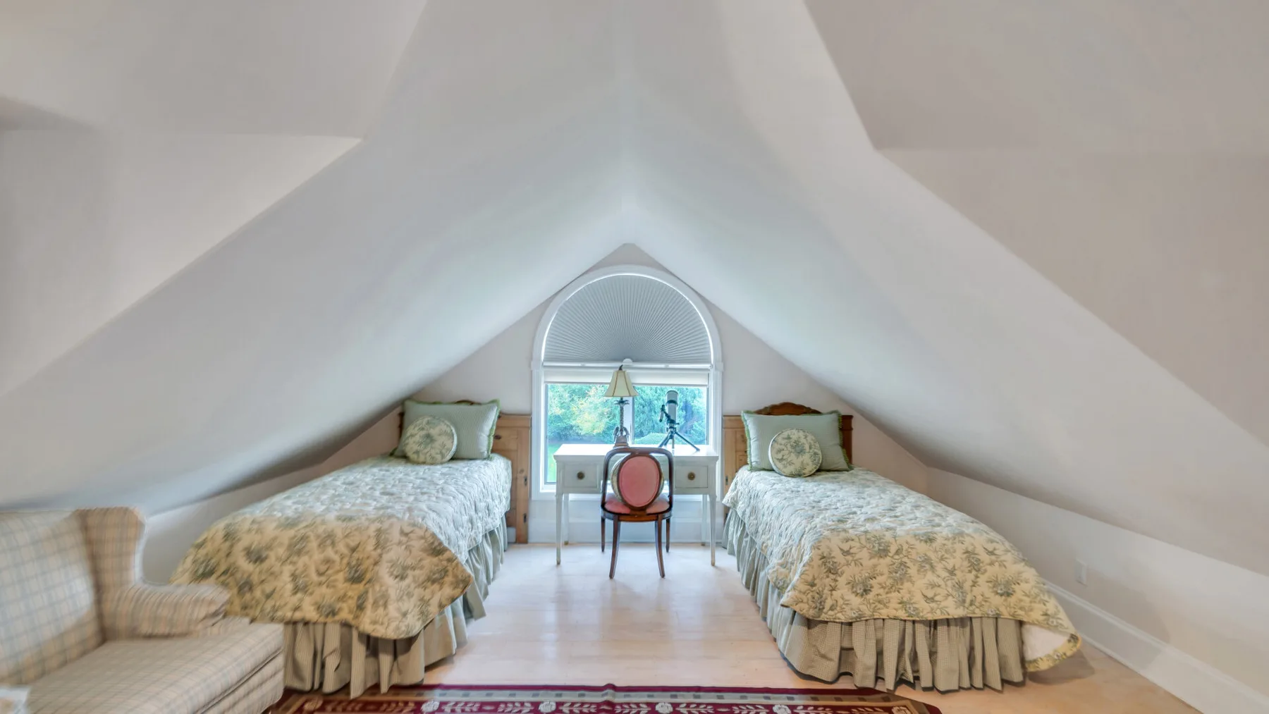 Un dormitorio abuhardillado con dos camas individuales y un escritorio bajo una ventana arqueada.