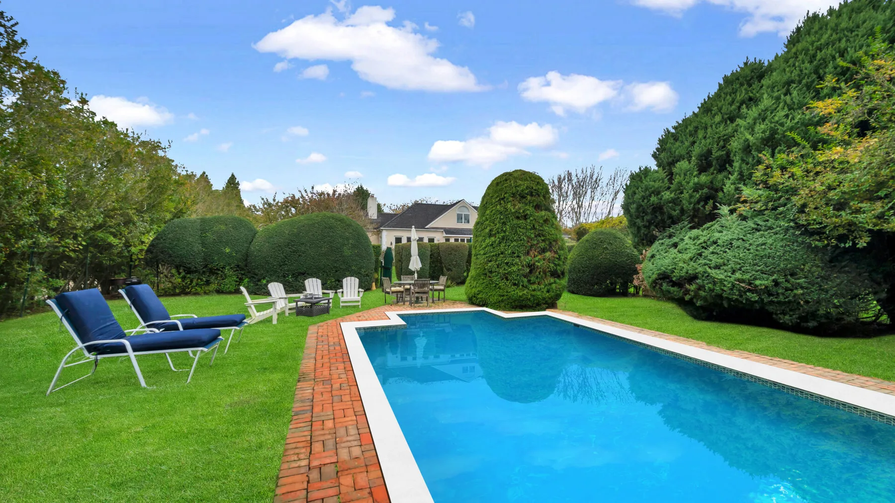 An outdoor swimming pool is surrounded by green lawn, seating, and hedges.