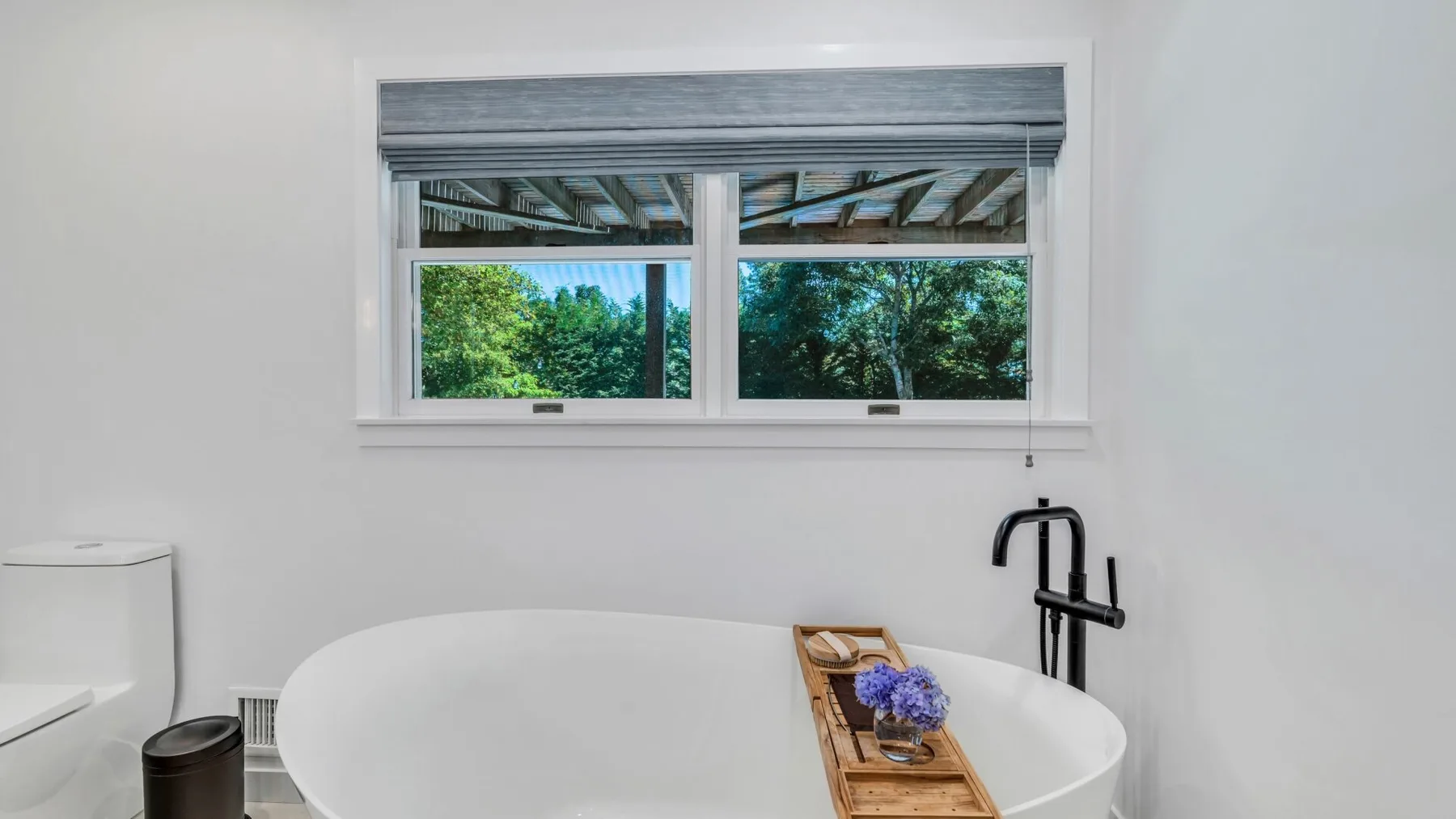 Un baño con una bañera blanca bajo una ventana con vista a la vegetación.