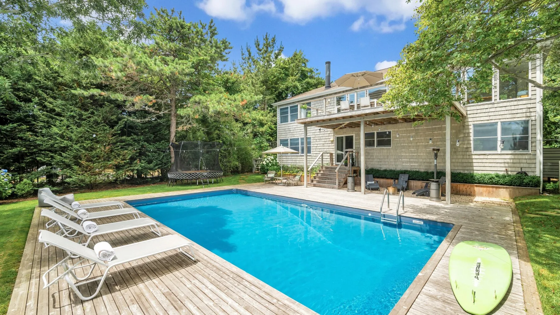 Una piscina en el patio está junto a una terraza de madera y una casa.