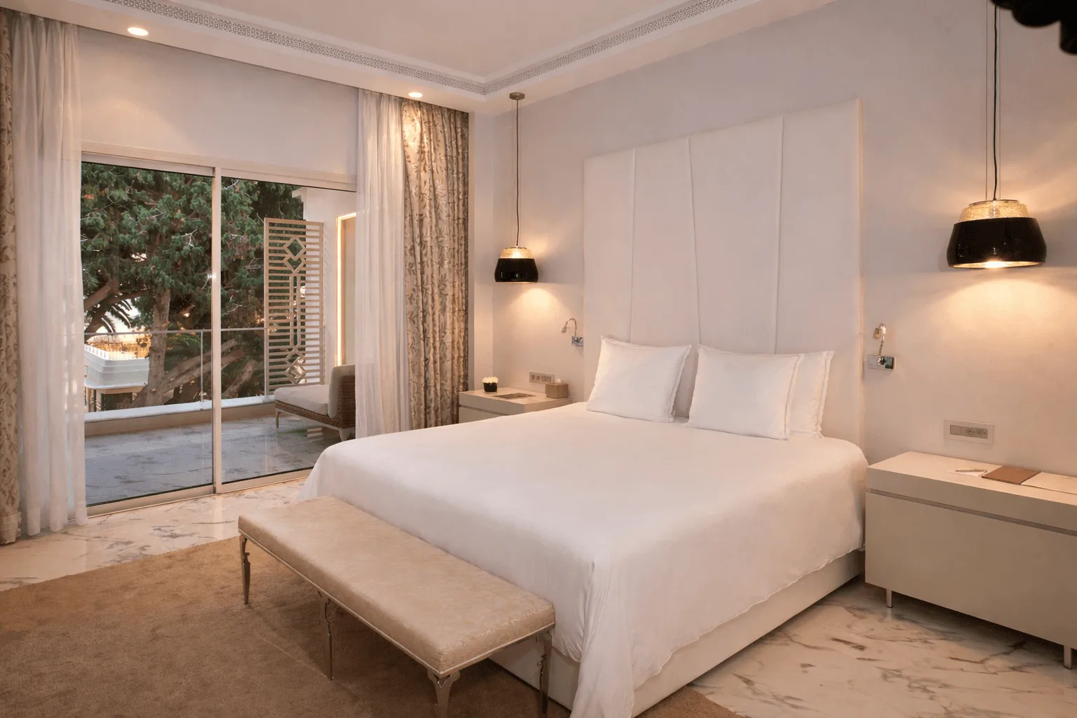 Suite de hotel moderna con una cama doble, un banco para sentarse y un gran ventanal que da a una terraza y a los árboles.