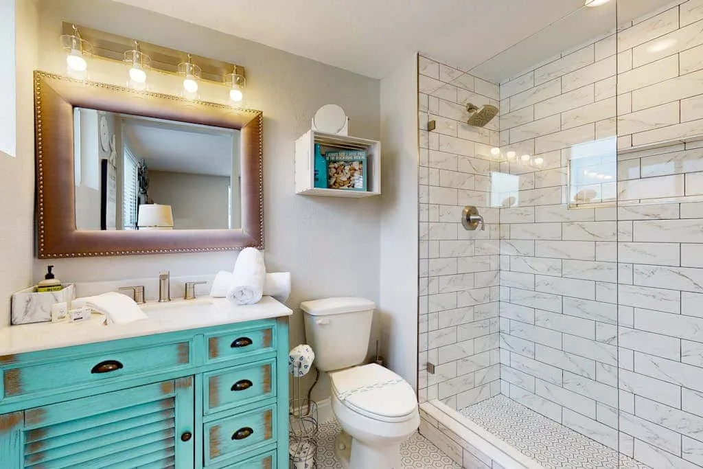 Un baño luminoso con un tocador turquesa, inodoro y ducha con azulejos.