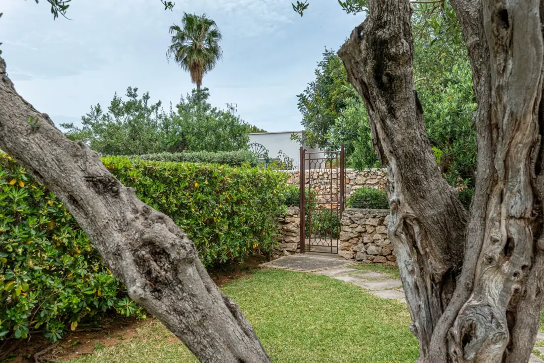 A garden path leads through a metal gate between stone walls and leafy hedges.