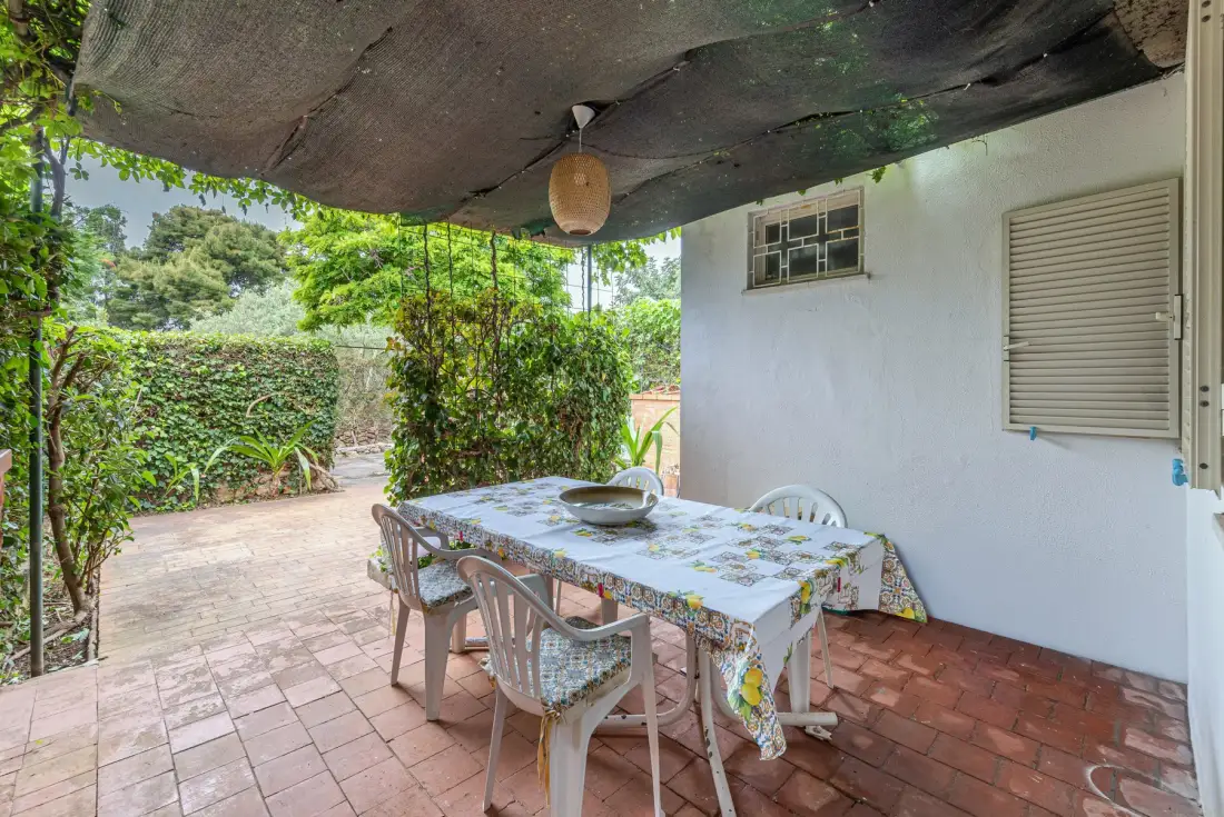 A shaded outdoor dining area has a table and seating beside a white wall.