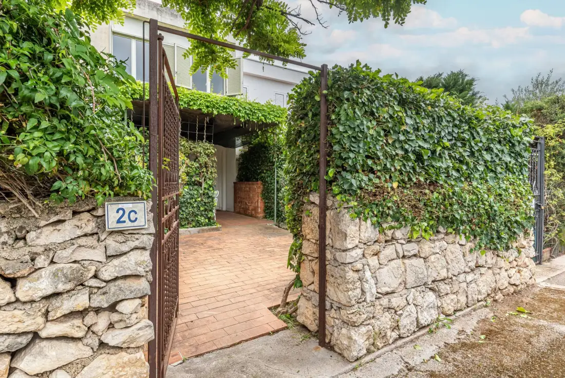 A brick-paved entrance passes through an open gate beside stone walls and greenery.