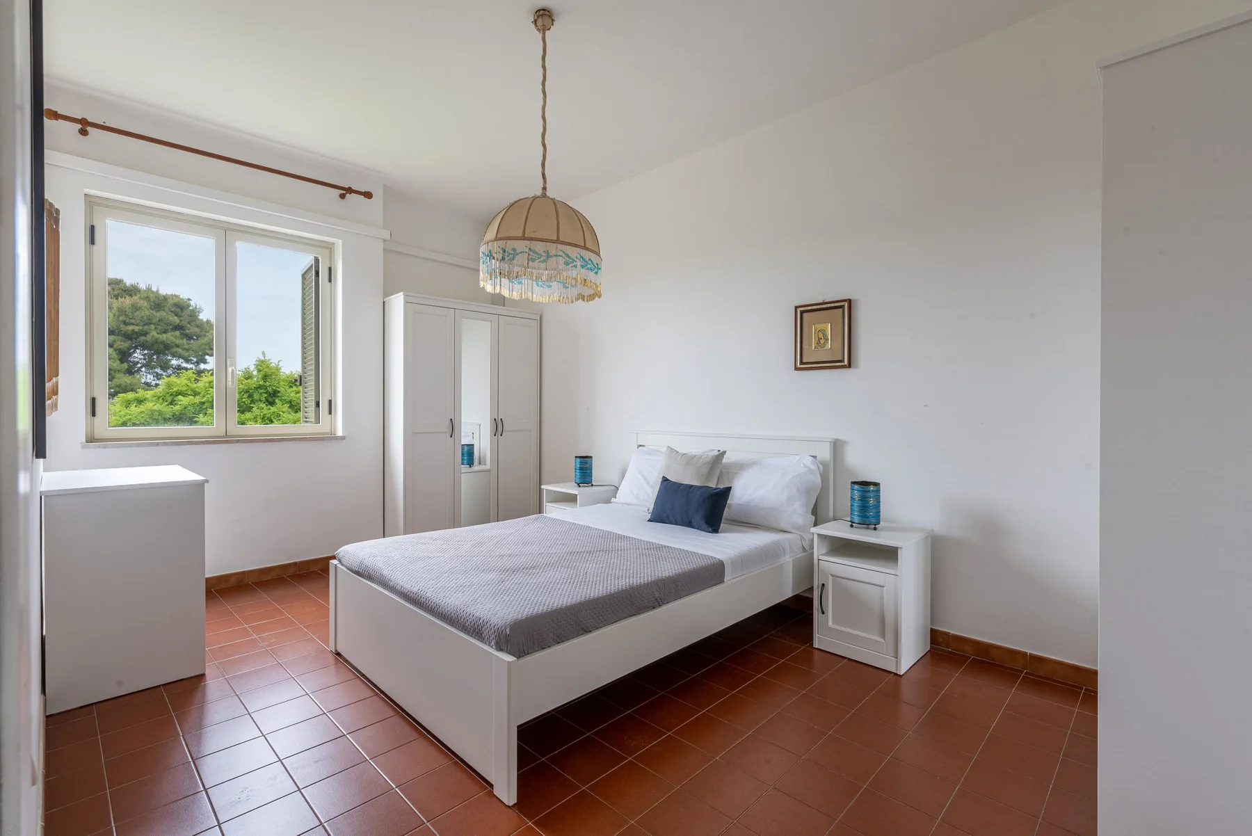A bright bedroom with a double bed, bedside tables, and a window overlooking greenery.