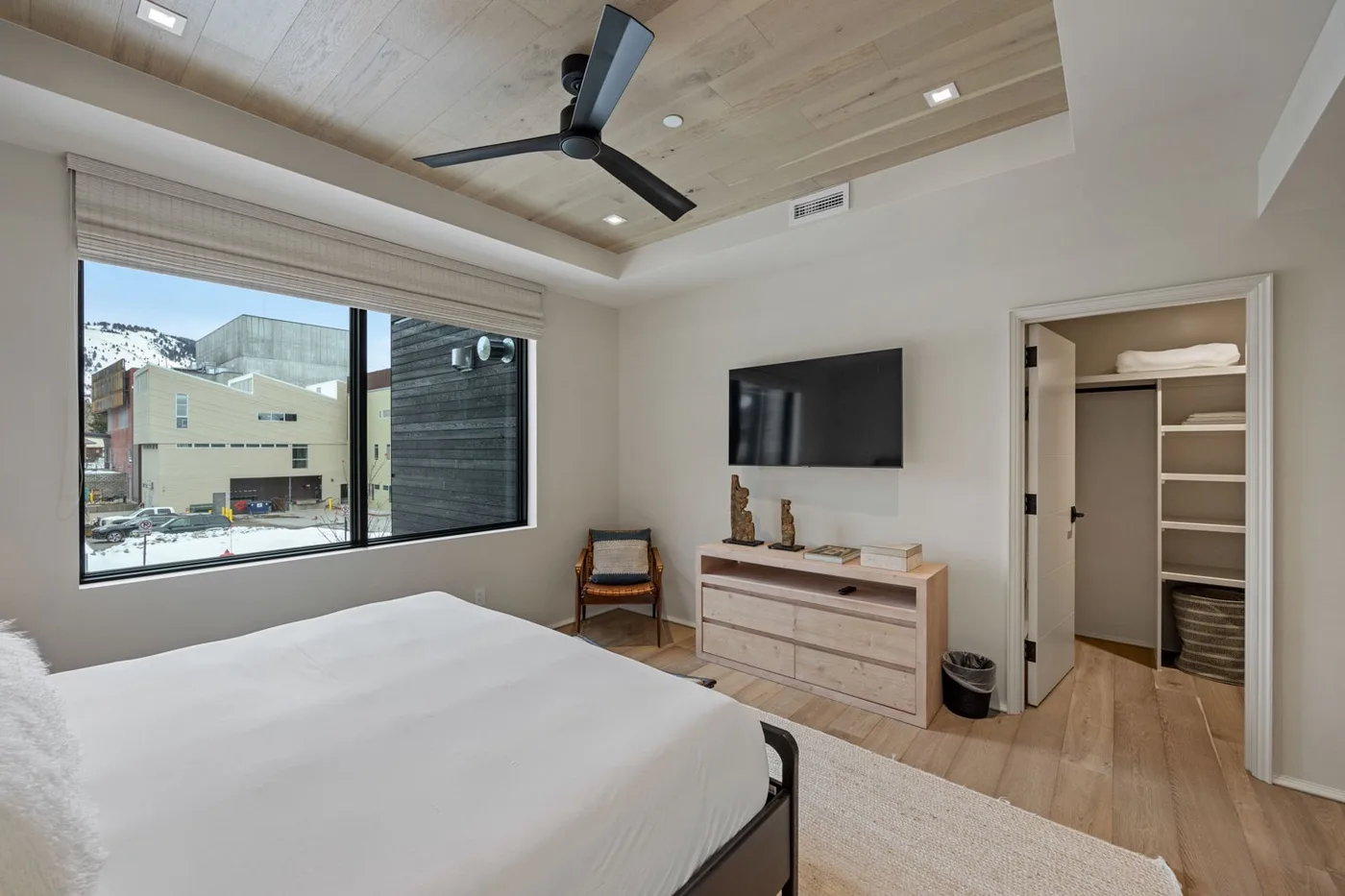 A bedroom with a bed, a wall-mounted television, and a window overlooking nearby buildings.