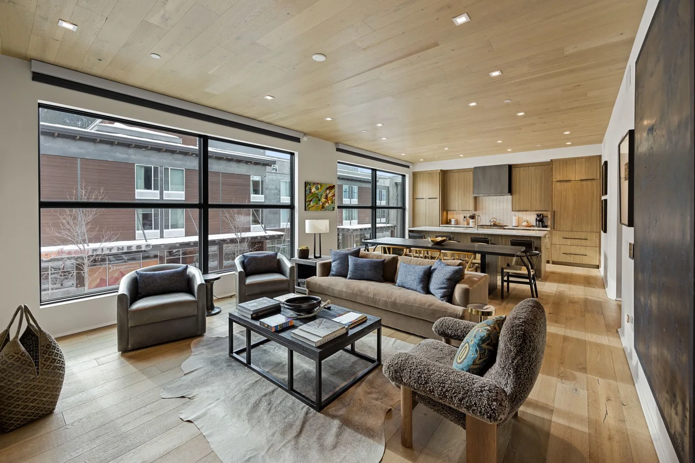 A living room with seating and large windows opens onto a kitchen and dining area.