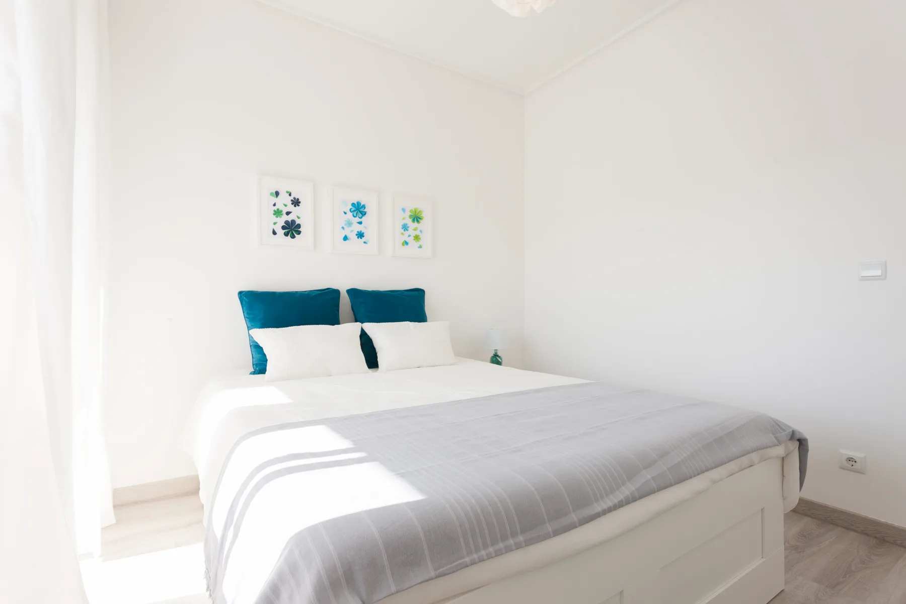 Un dormitorio luminoso con una cama, almohadas turquesas y pequeños cuadros en la pared.