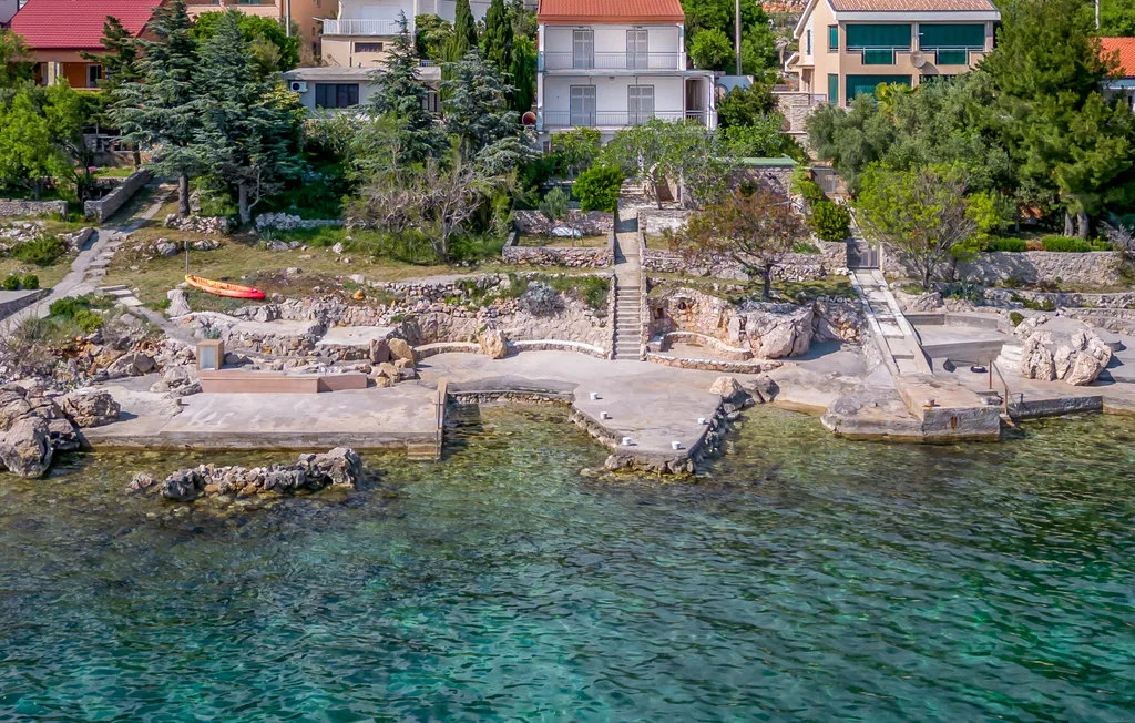 A rocky waterfront with clear water, stone steps, and houses among trees.