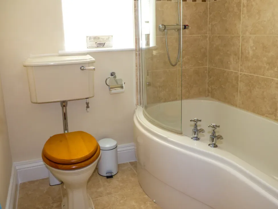 A bathroom with a toilet with wooden seat a white bin and a bathtub with tiled walls at Lockton Cottage in Pickering
