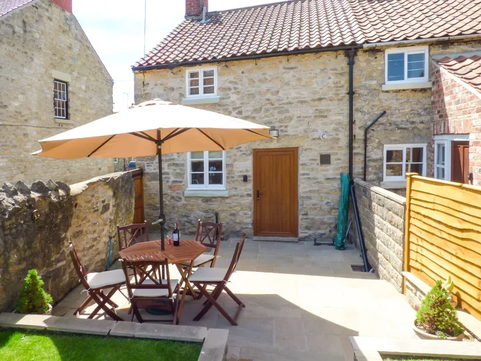 An outdoor patio with a wooden table and four chairs under a beige umbrella in front of a stone house at Lockton Cottage in Pickering