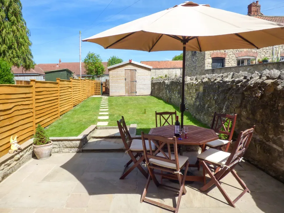 An outdoor patio area with a wooden table and chairs under a large umbrella next to a stone wall and a garden shed at Lockton Cottage in Pickering