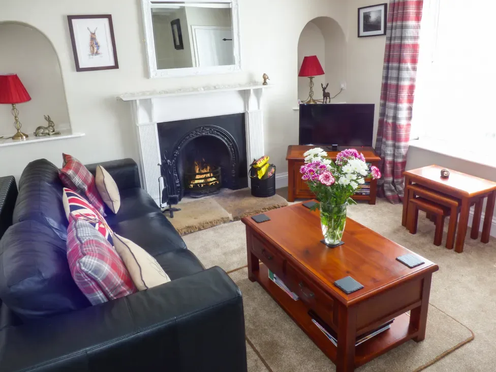 A living room with a black sofa and cushions a fireplace with a fire a wooden coffee table with flowers a television and matching side tables at Lockton Cottage in Pickering