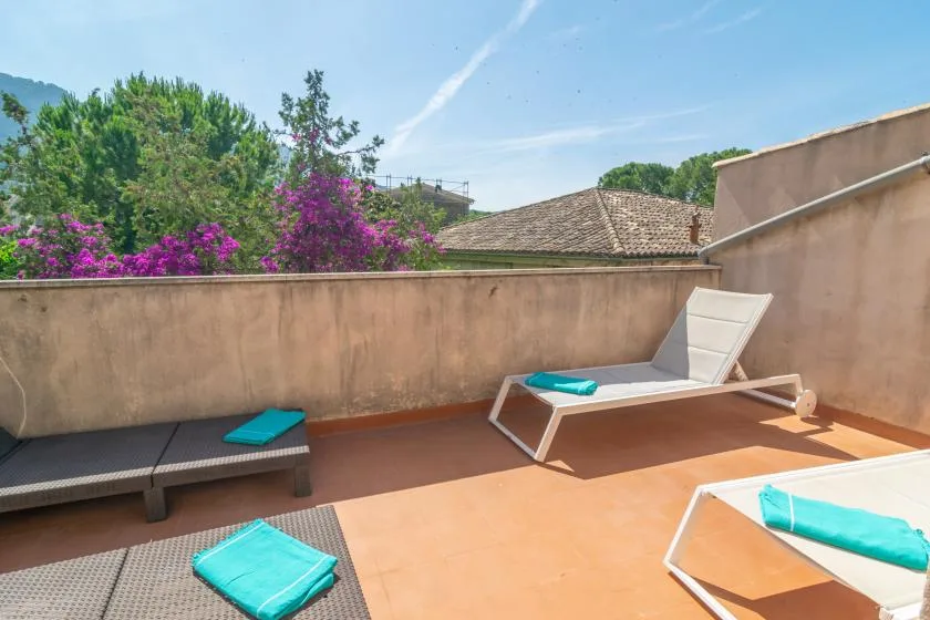A sunny terrace with loungers, turquoise cushions, and flowering greenery beyond the wall.