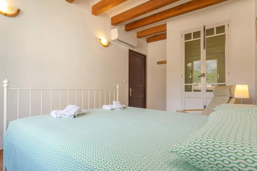 A bedroom with a large bed, pale patterned bedding, and exposed wooden ceiling beams.