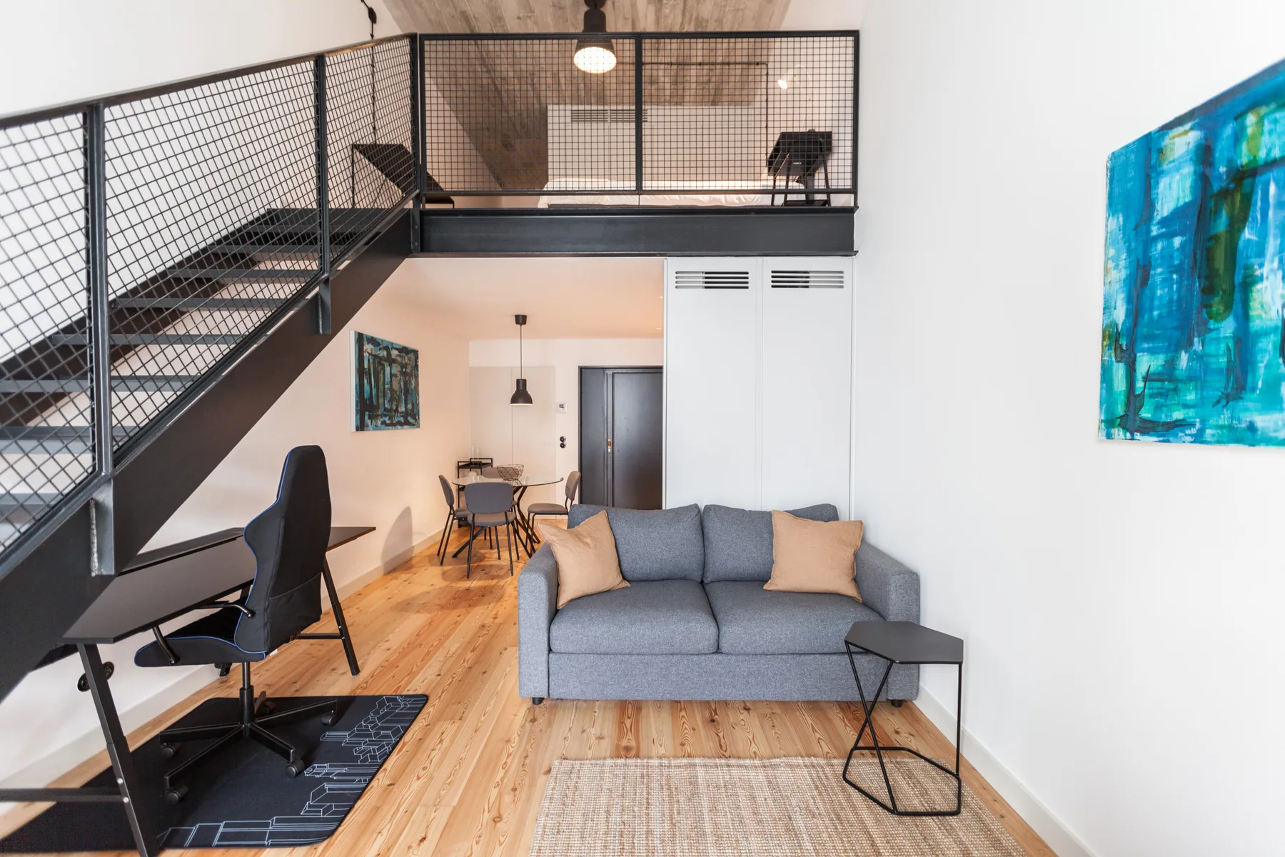 Sala de estar con asientos bajo un altillo con barandilla metálica y escaleras.