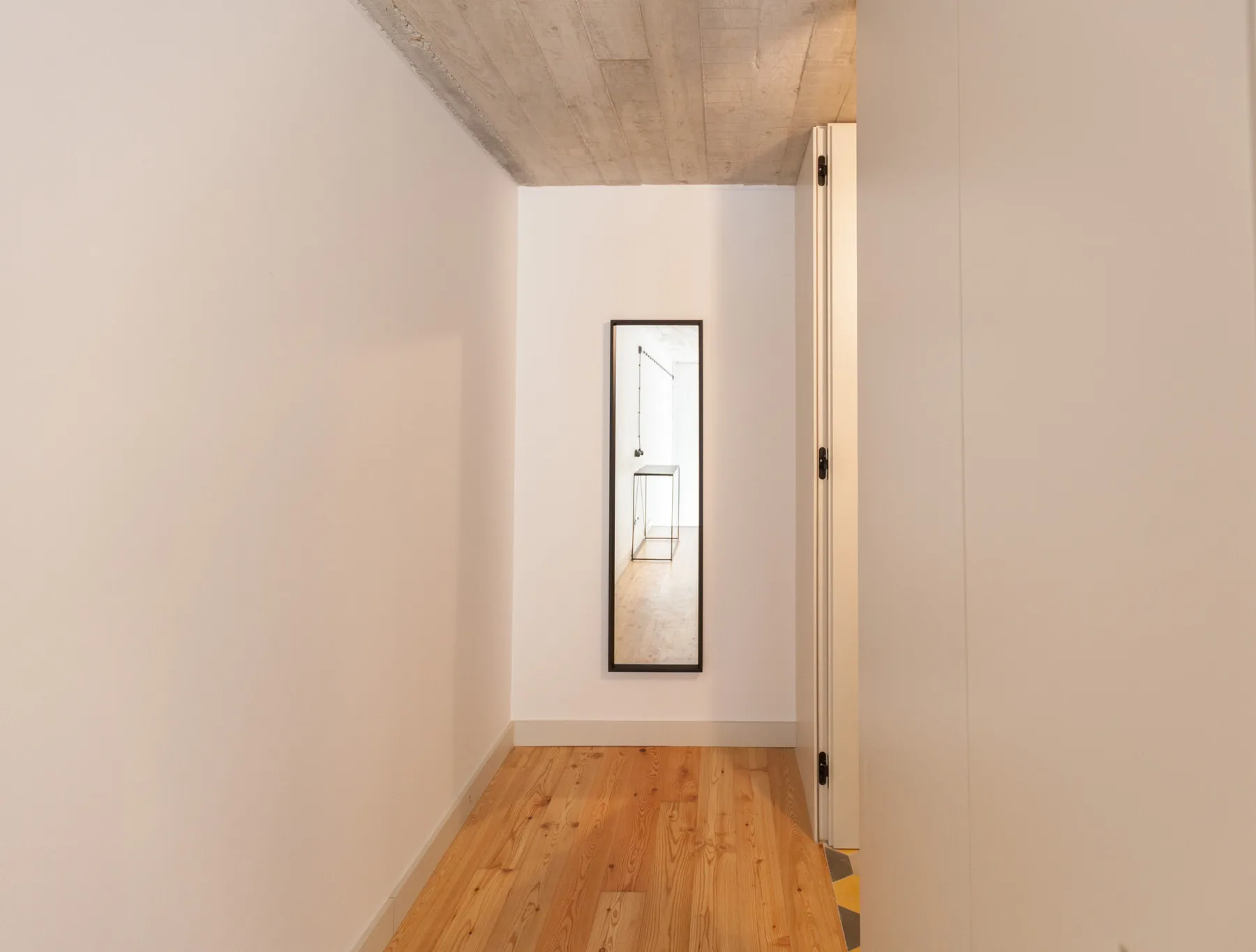 A narrow hallway with light wood flooring and a framed mirror on the wall.