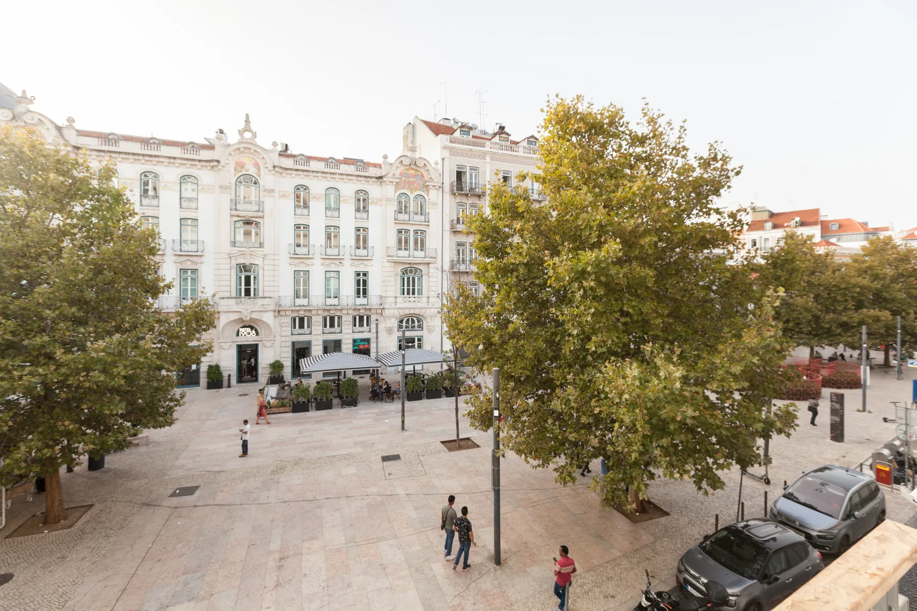 A paved urban plaza with trees, pedestrians, and surrounding buildings.