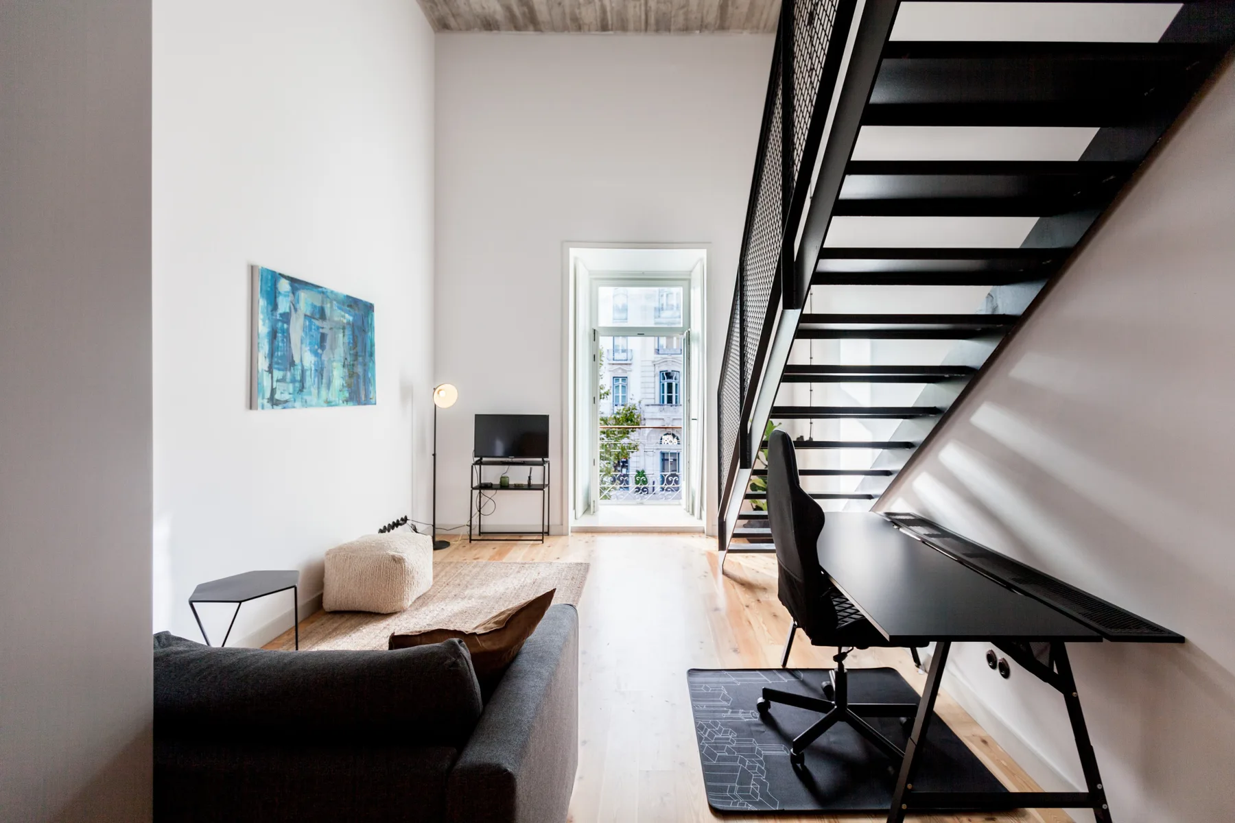 A bright living area has seating, a desk, and an open staircase.