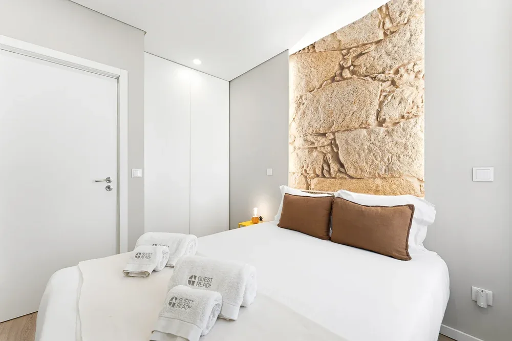 A bright bedroom with a white bed and a textured stone wall behind it.
