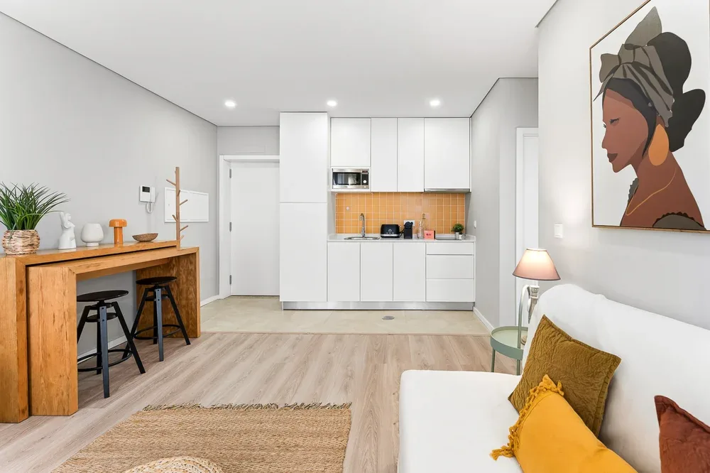 A bright open-plan room with a wooden desk, kitchenette, and seating.