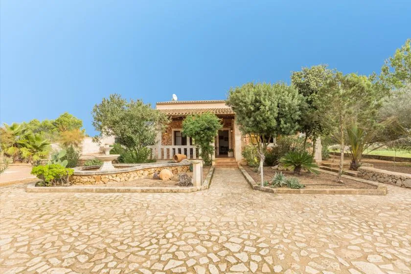 A low house stands behind a broad stone-paved courtyard with trees and planted areas.