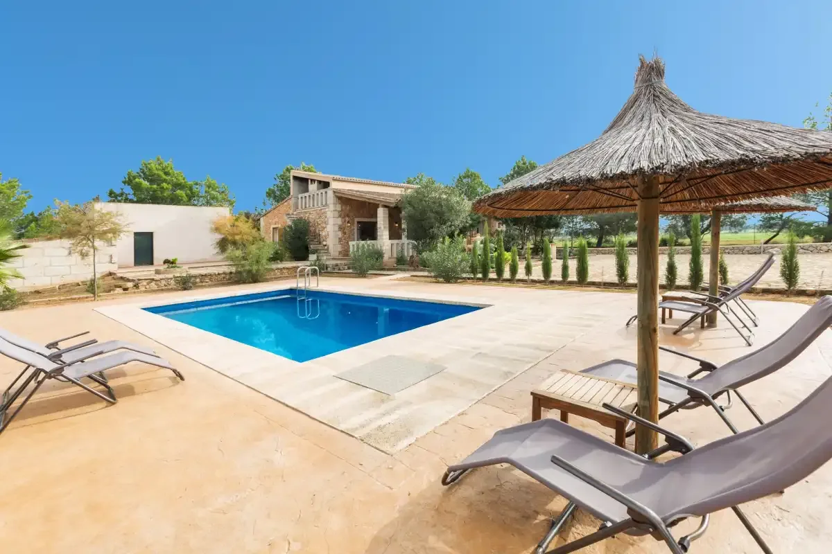 An outdoor swimming pool sits beside a paved patio with reclining seats and a thatched parasol.