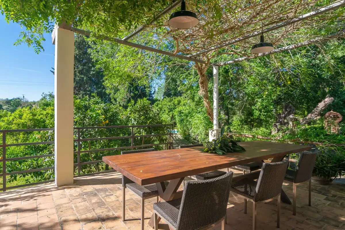 Una zona de comedor exterior sombreada tiene una mesa larga bajo una pérgola frondosa.