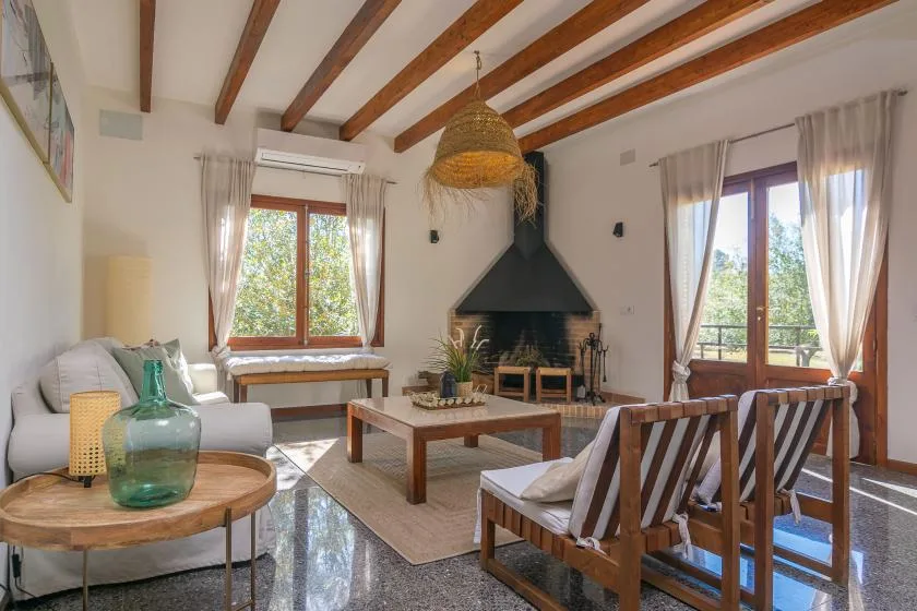 Una sala luminosa con chimenea, asientos de madera y grandes ventanas con cortinas.