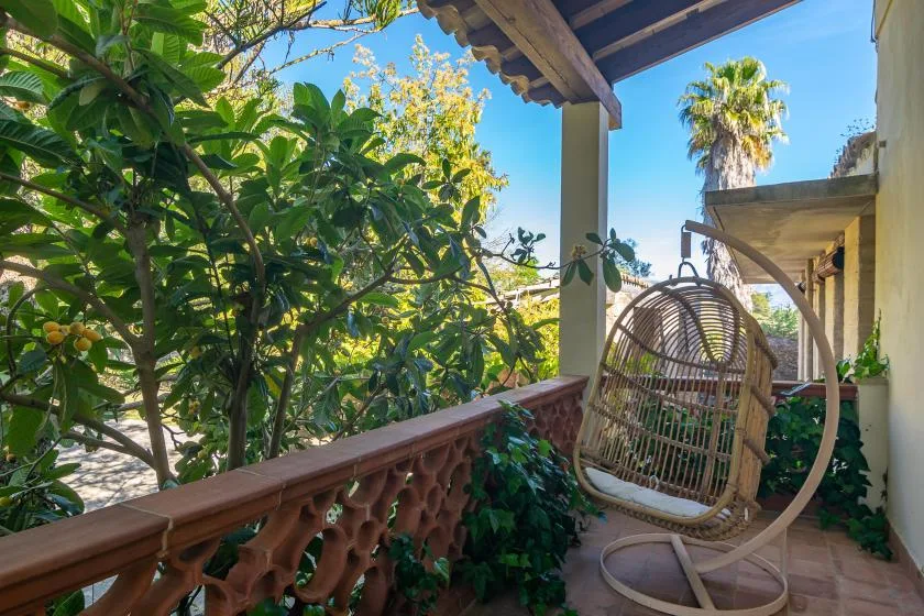 Un balcón sombreado con una silla colgante frente a árboles frondosos.