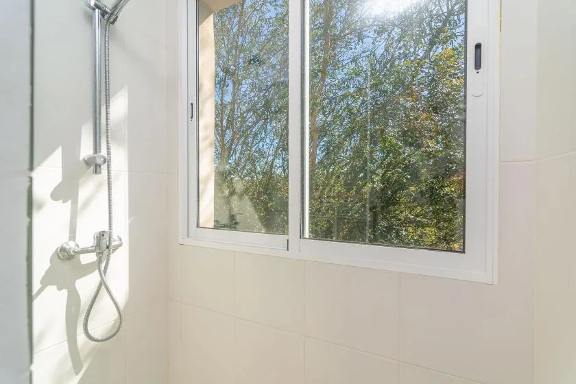 Un baño luminoso con una ducha de pared junto a una ventana grande con vista a los árboles.